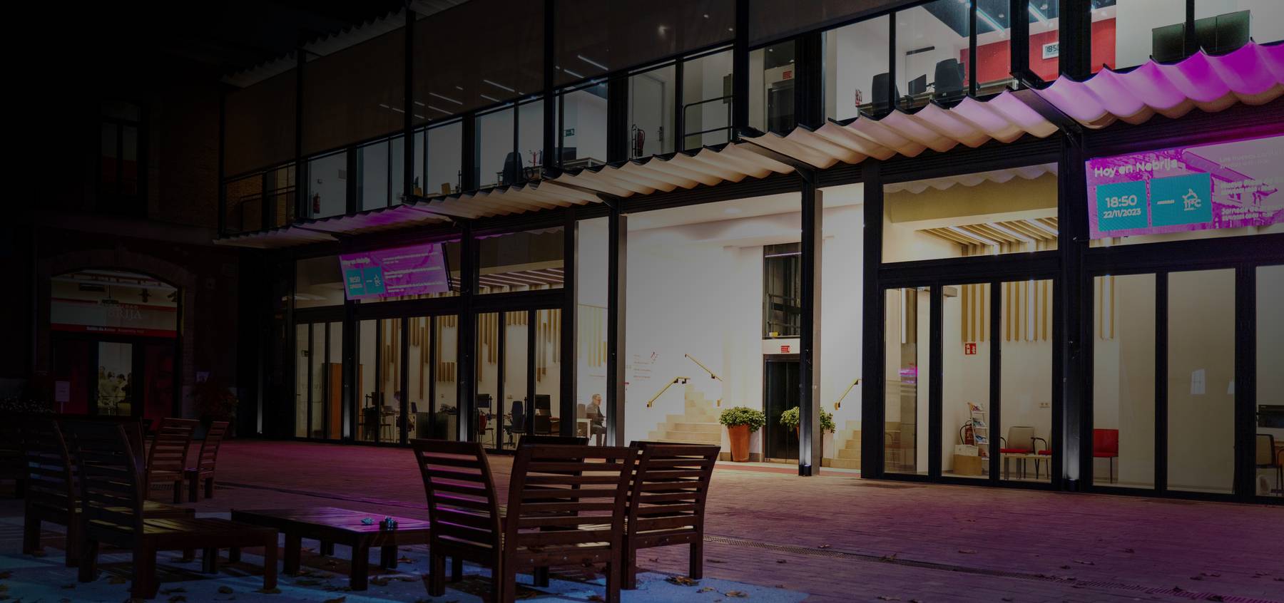 Una fachada moderna con sillas de madera en un ambiente nocturno y luces de colores.