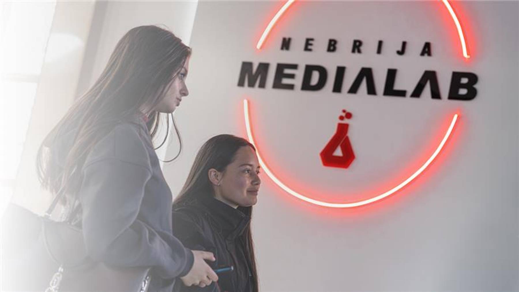 Dos jóvenes se encuentran en un espacio moderno con un letrero iluminado que dice 'NEBRIJA MEDIALAB'.