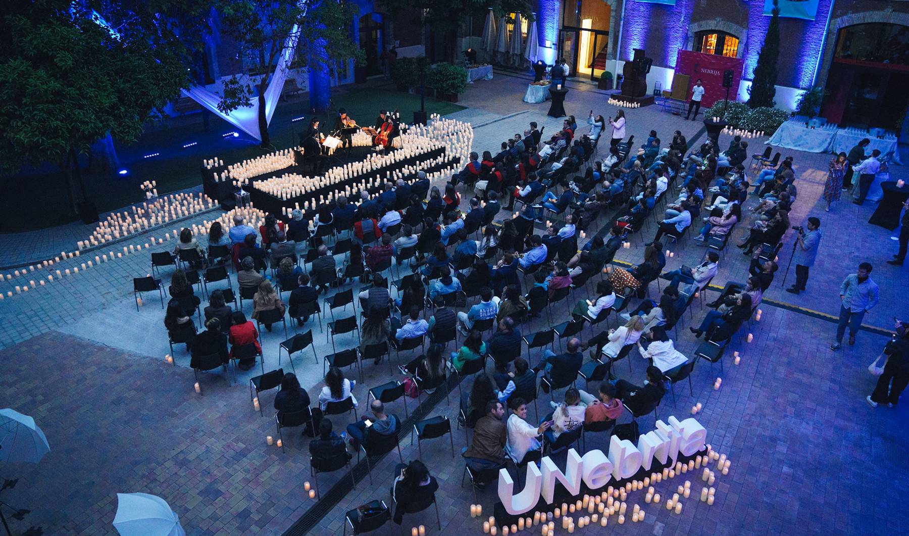 Un evento al aire libre con un gran número de asistentes sentados en sillas rodeados de velas y decoraciones iluminadas.