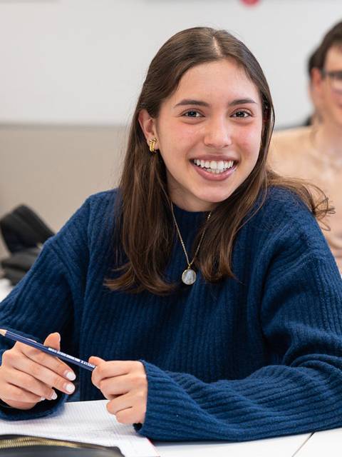 Una joven sonriente en un aula, sosteniendo un bolígrafo y mirando hacia la cámara.
