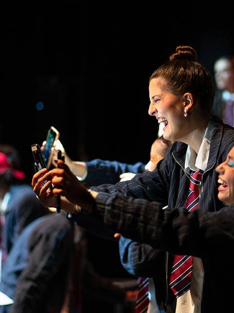 Un grupo de jóvenes en un escenario se ríen y se toman selfies con sus teléfonos móviles.