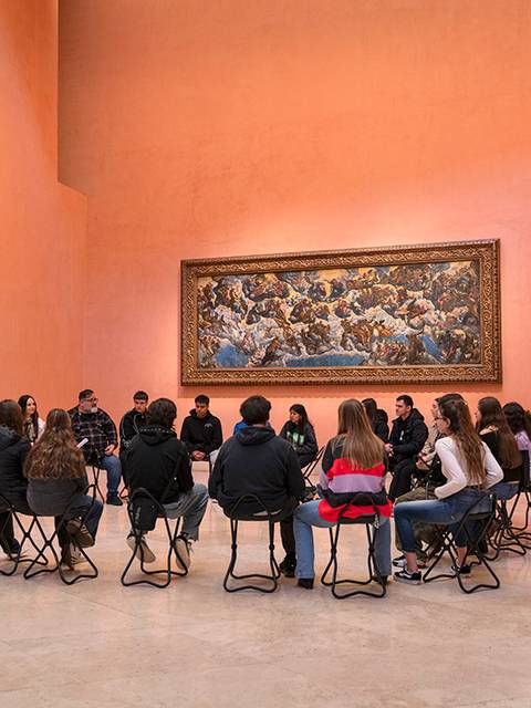 Un grupo de personas sentadas en círculo en una sala de museo con paredes de color melocotón y una gran pintura en la pared.