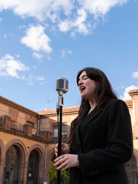 Una joven canta frente a un micrófono en un patio con arquitectura histórica bajo un cielo azul.