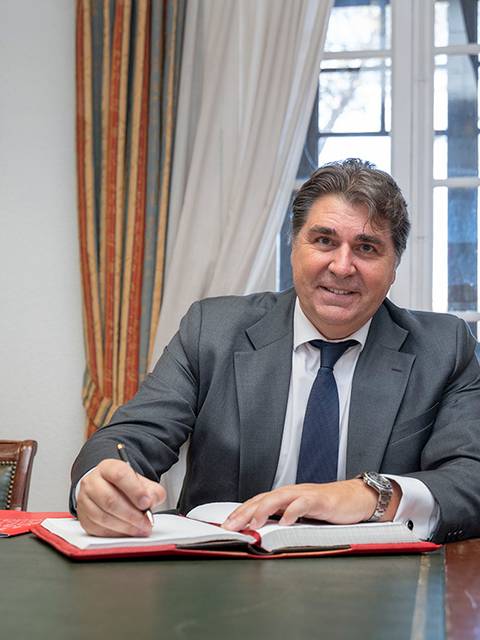 Un hombre sonriente, vestido con traje, está sentado a una mesa escribiendo en un libro en un ambiente interior iluminado.