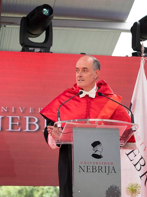Un hombre con toga habla en un podio frente a un fondo con el nombre de la Universidad Nebrija.