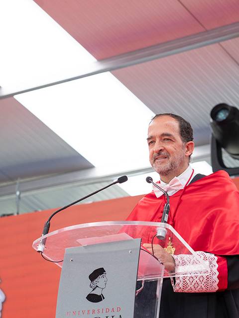 Un hombre con toga académica habla desde un podio en un evento formal.