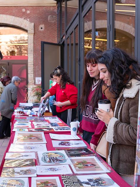 Un grupo de mujeres jóvenes observa una mesa con carteles y productos en un evento al aire libre.