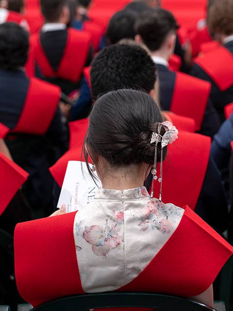 Un grupo de personas sentadas con capas rojas, enfocándose en una mujer que lleva un vestido con detalles florales.