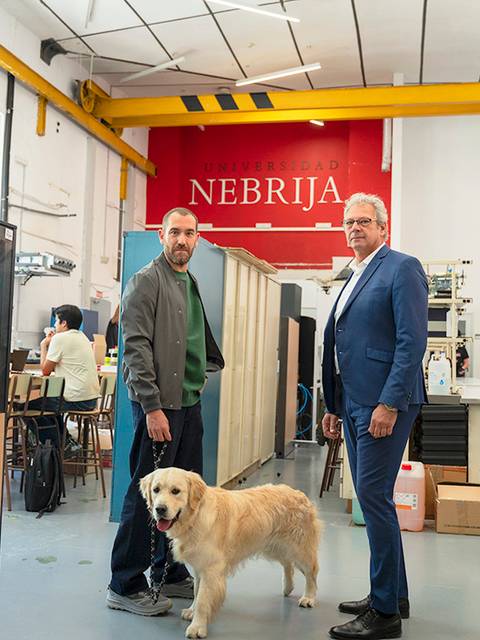 Dos hombres de pie en un laboratorio o taller, uno con un perro, bajo un letrero que dice 'NEBRIJA'.