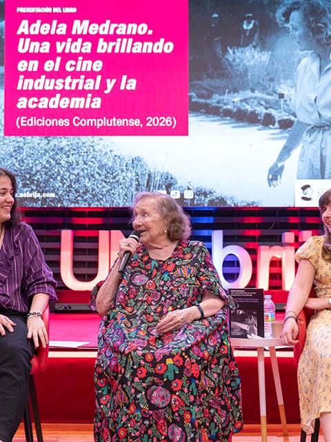 Una charla en un evento cultural con tres mujeres sentadas frente a una presentación sobre Adela Medrano.