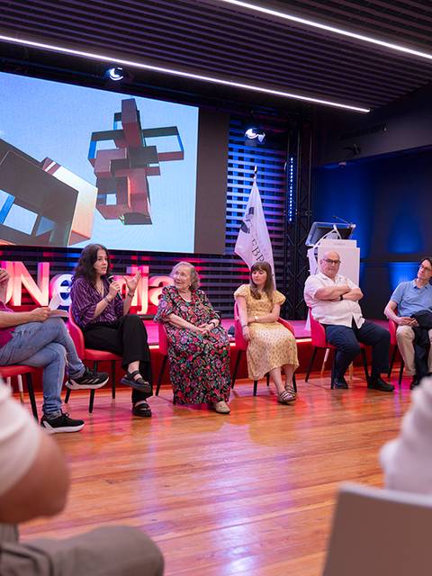 Un grupo de personas discute en un evento mientras una proyección se muestra en la pantalla al fondo.
