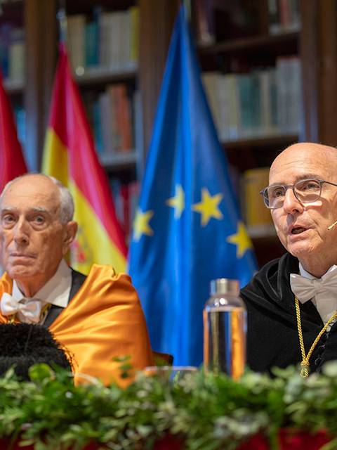 Dos hombres adultos en ceremonia formal, sentados frente a banderas de España y la Unión Europea.