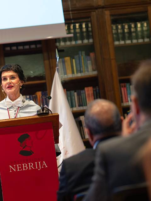 Una mujer habla en un podio durante un evento académico en una biblioteca.