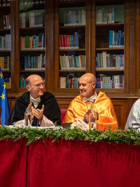 Cuatro personas ataviadas con togas en una ceremonia académica, rodeadas de banderas y estanterías con libros.