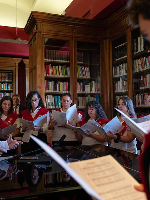 Un grupo de personas cantando en una biblioteca mientras un director observa atentamente.