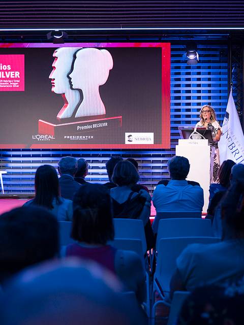 La imagen muestra a una mujer hablando en un escenario durante la entrega de los premios Belsilver, con un gran pantalla detrás y asistentes en primer plano.