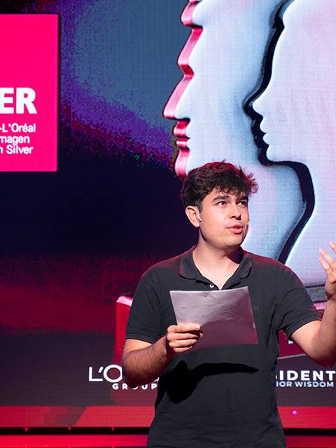 Un joven presenta los Premios Belsilver en un escenario con un fondo colorido.