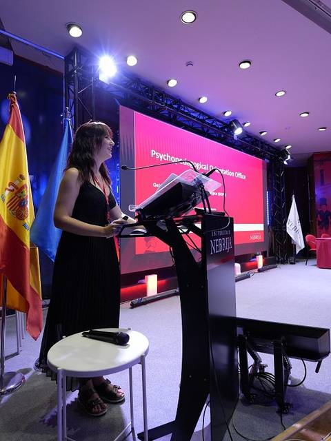 Una presentadora habla en un evento con banderas y una pantalla de fondo.