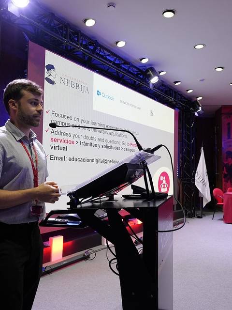 Un hombre está de pie frente a un podio en una sala de conferencias, mientras presenta información sobre servicios educativos.