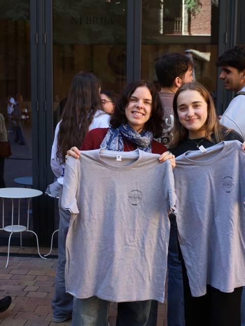 Dos chicas sostienen camisetas grises frente a un grupo de personas en un evento.