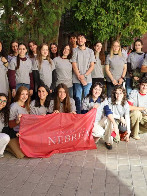 Un grupo de estudiantes posando juntos, algunos con camisetas grises, sosteniendo una bandera del Club Nebrija en un evento de diseño.
