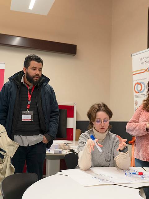 Un grupo de cuatro personas interactúa en una reunión, con una mujer escribiendo en un papel en una mesa.