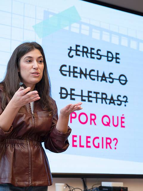 Una mujer habla frente a una pantalla con un mensaje sobre la elección entre ciencias y letras.