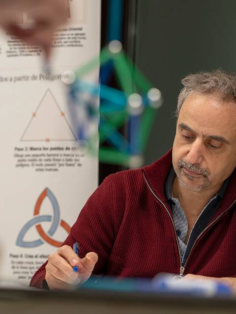 Un hombre y una mujer están trabajando juntos en una actividad relacionada con geometría, con varios modelos tridimensionales en el fondo.