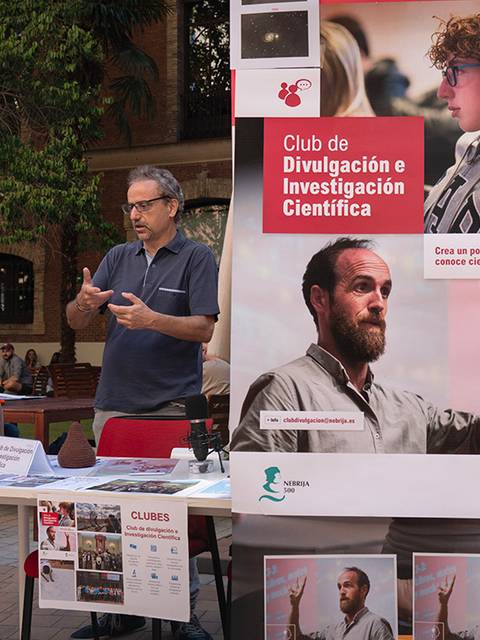 Dos hombres conversan frente a un stand sobre divulgación e investigación científica en un ambiente al aire libre.