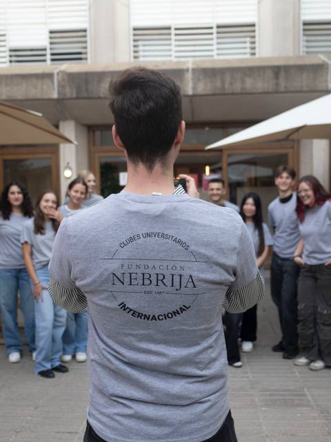 Un grupo de estudiantes posa al aire libre frente a un edificio, mientras uno de ellos les toma una foto desde atrás.