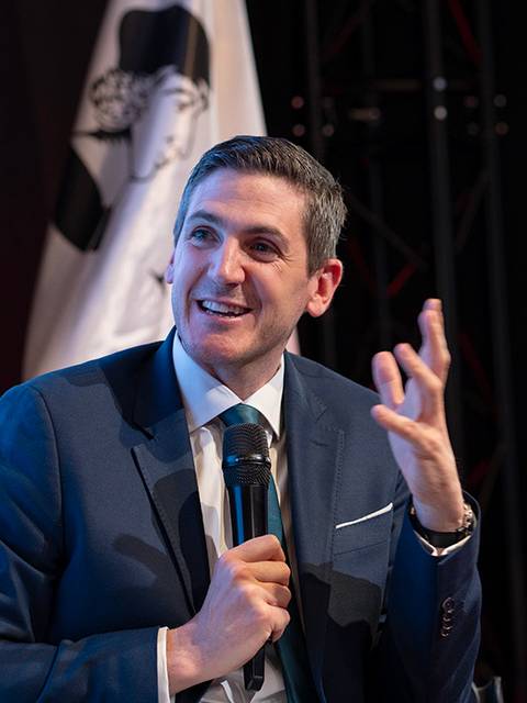 Un hombre sonriente habla en un evento con un micrófono en la mano.