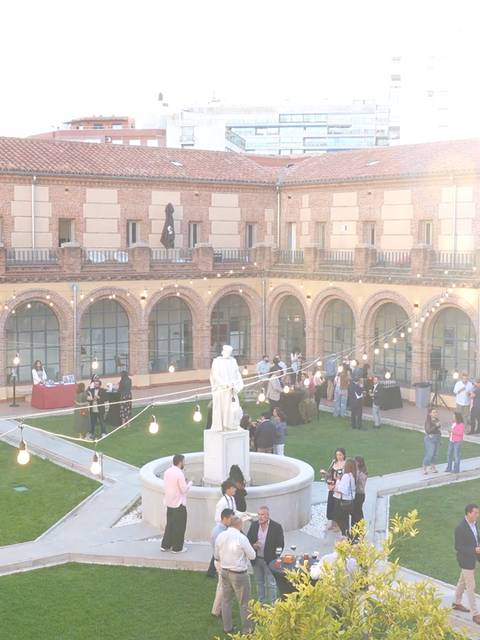 La imagen muestra un evento al aire libre en un patio con una estatua en el centro y personas conversando y socializando alrededor.