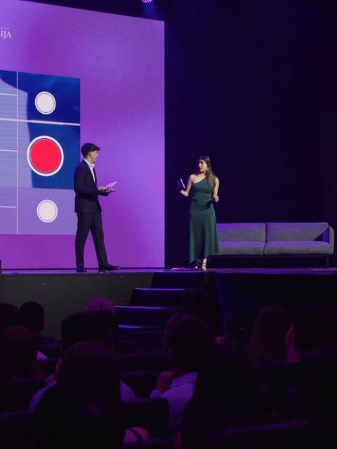Un hombre y una mujer están en el escenario de un evento, interactuando frente a una presentación visual con elementos gráficos.