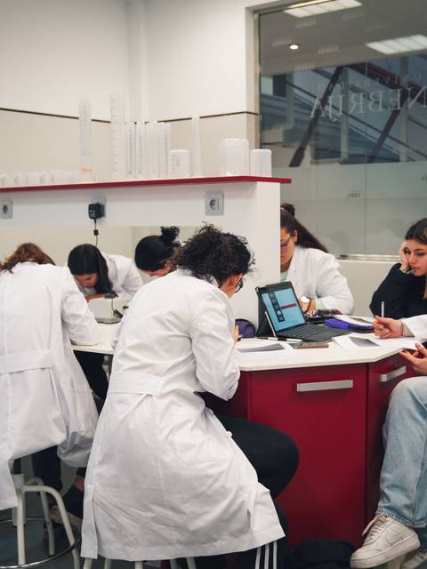 Un grupo de estudiantes en un laboratorio, todos con batas blancas, trabajando en sus tareas en una mesa.