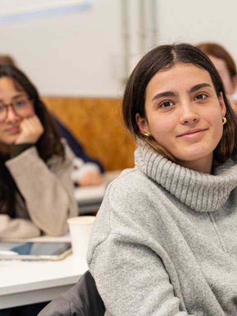 Una estudiante sonriente en un aula, con compañeras al fondo atendiendo a la clase.