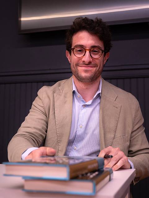 Un hombre sonriente sentado en una mesa con libros delante de él.