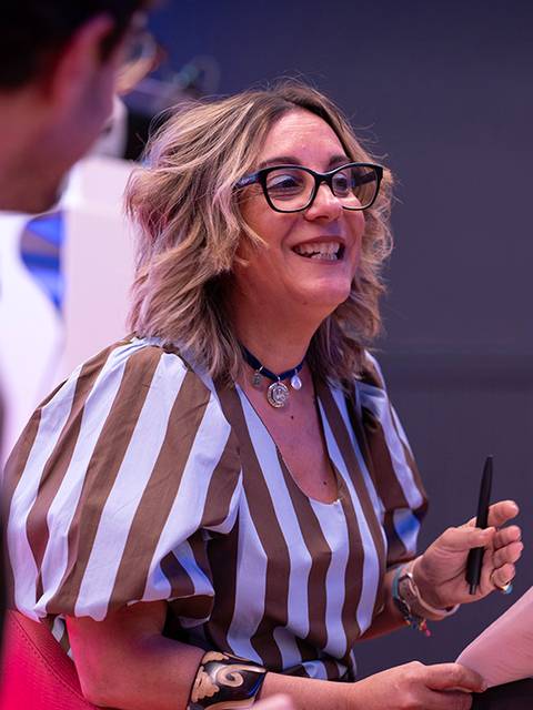 Una mujer sonriente con gafas sostiene papeles mientras habla, vestida con una blusa de rayas.