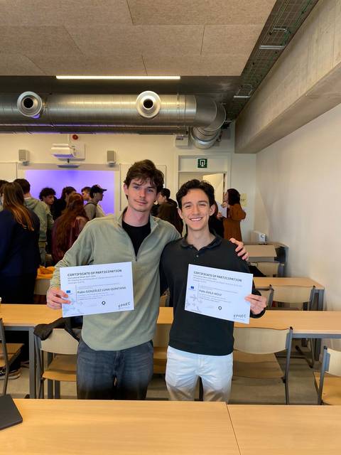 Dos jóvenes sosteniendo certificados en un espacio con múltiples personas al fondo.