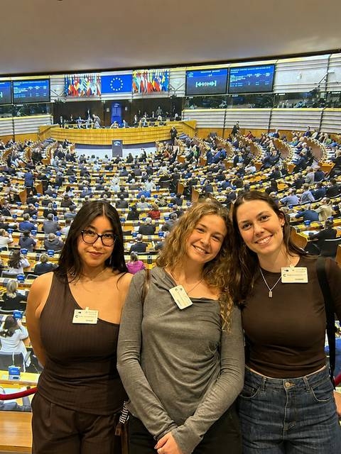 Tres jóvenes posan en un entorno del Parlamento Europeo con un gran auditorio al fondo.