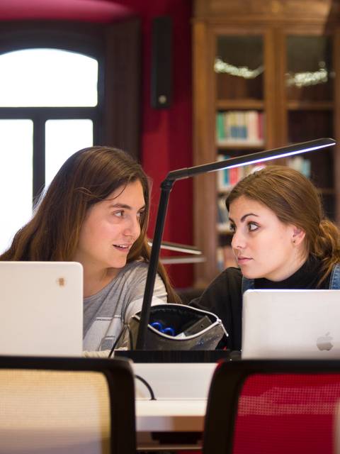 Dos jóvenes conversan en una biblioteca mientras trabajan en sus laptops.