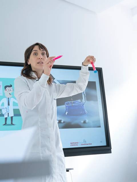 Una mujer con bata de laboratorio ofrece una presentación interactiva en un aula moderna.
