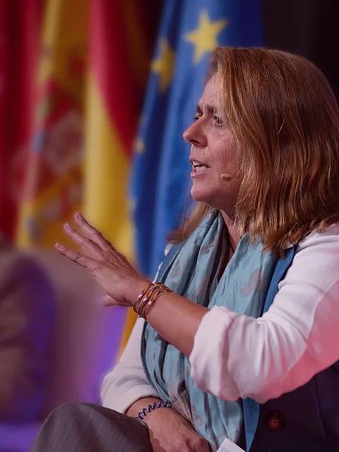 Una mujer está hablando en un evento mientras un hombre escucha al fondo.