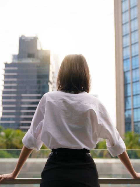 Una mujer observa la ciudad desde un balcón con rascacielos al fondo.