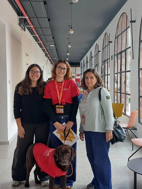 Tres personas posan en un vestíbulo moderno junto a un perro labrador con un abrigo rojo.