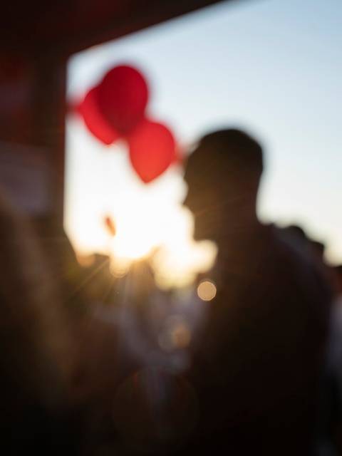 La imagen muestra un grupo de personas en un entorno festivo al atardecer, con globos rojos en el fondo.