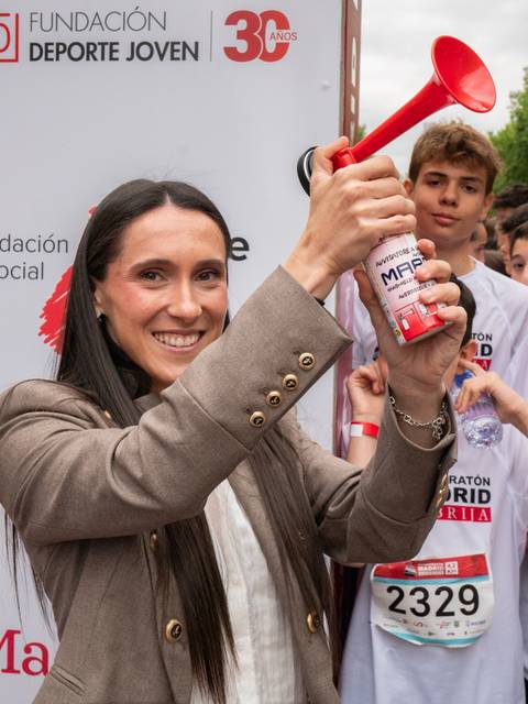 Una mujer sostiene un megáfono rojo mientras sonríe en un evento deportivo con jóvenes al fondo.