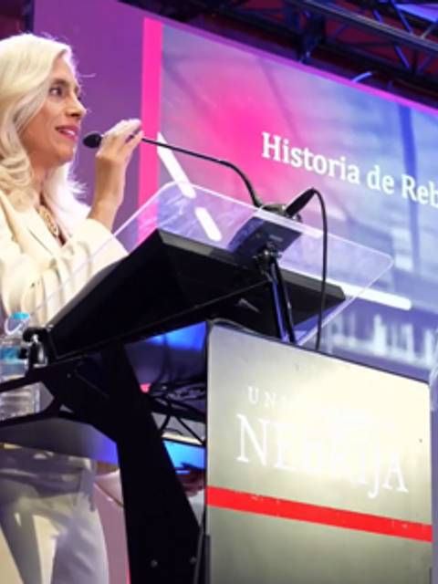 Una mujer presenta un evento en un auditorio con pantallas que muestran información sobre 'Historia de Rebeca'.