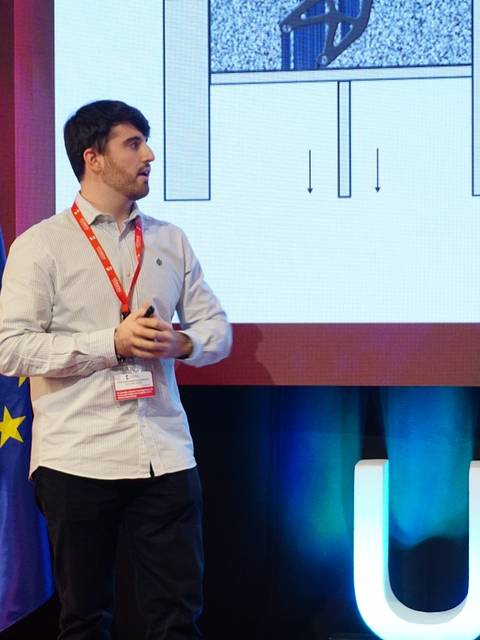 Un hombre presenta un proyecto en un escenario con banderas de España y la Unión Europea de fondo.