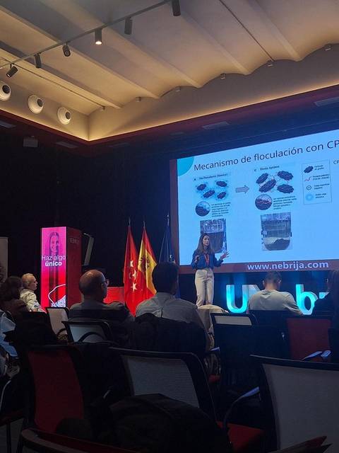 Una presentadora expone sobre el mecanismo de floculación en un evento, con un público atento y banderas de fondo.