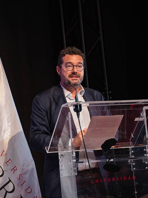 Un hombre de pie frente a un podio con una bandera de la Universidad Nebrija al fondo.